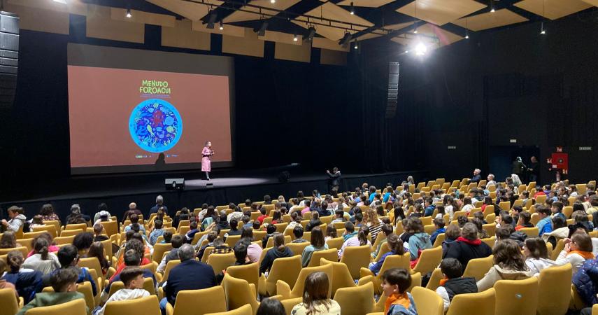 Apertura de MenudoForoAcui. Auditorio cheo de crianzas, coa presentadora no escenario e a imaxe do proxecto na pantalla do fondo.